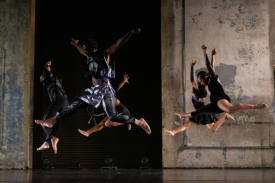 6 dancers leap through the air in front of a concrete wall.